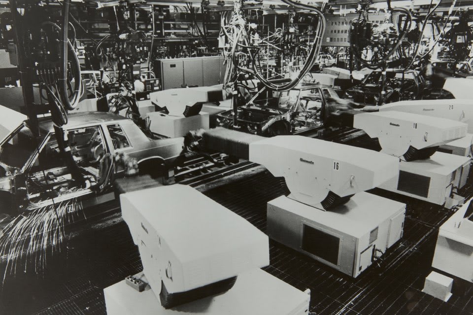 Crno-bijela fotografija tvorničke montažne linije s nekoliko robota Unimate iz 1960-ih i nedovršenih karoserija automobila, od kojih se jedna zavaruje i iskre, dok se iznad vide cijevi i kablovi.